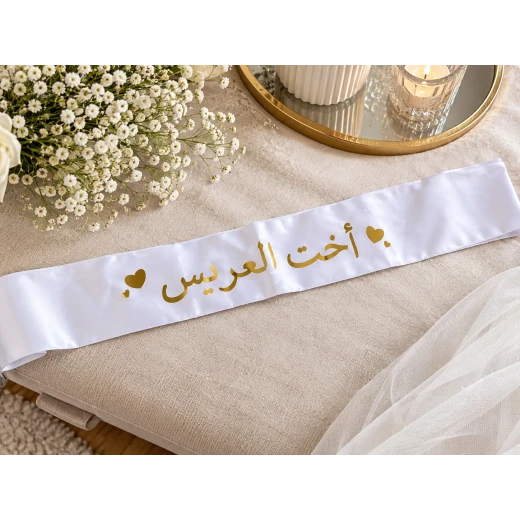 White Satin Wedding Sash with Gold “Groom’s Sister” Arabic Lettering and Heart Details White Satin Wedding Sash with Gold “Groom’s Sister” Arabic Lettering and Heart Details